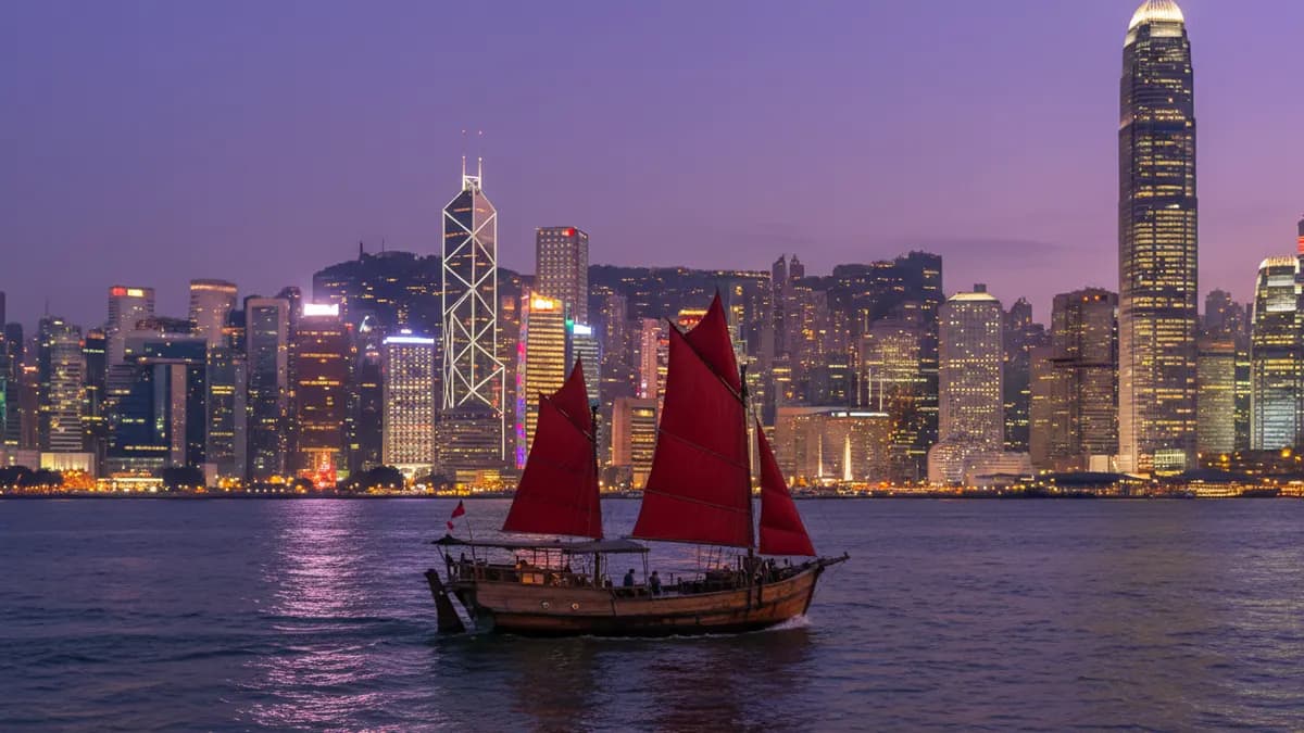 A harbour city skyline at dusk with a red-sailed boat on the water
