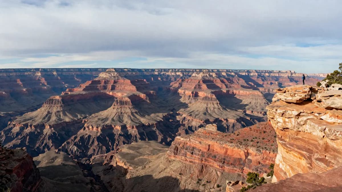 A vast canyon with layered red rock walls and a lone hiker on the rim